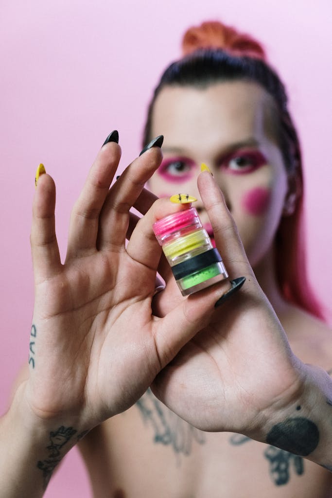 Hands holding vibrant cosmetic products with a tattooed model in pink makeup.